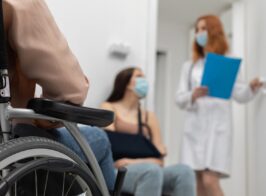 In The Foreground A Wheelchair And In The Background A Redheaded Doctor Standing Next To Her Doctor's Office. A Protective Mask Over Her Mouth And Nose.